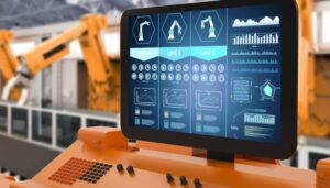 Immagine di un monitor che mostra un'interfaccia uomo-macchina in un ambiente industriale. L'interfaccia permette il controllo e il monitoraggio delle linee di produzione, rappresentando l'uso di tecnologie avanzate e automatizzate nelle imprese per migliorare l'efficienza operativa. Sullo sfondo sono visibili bracci robotici che operano nella catena di montaggio.